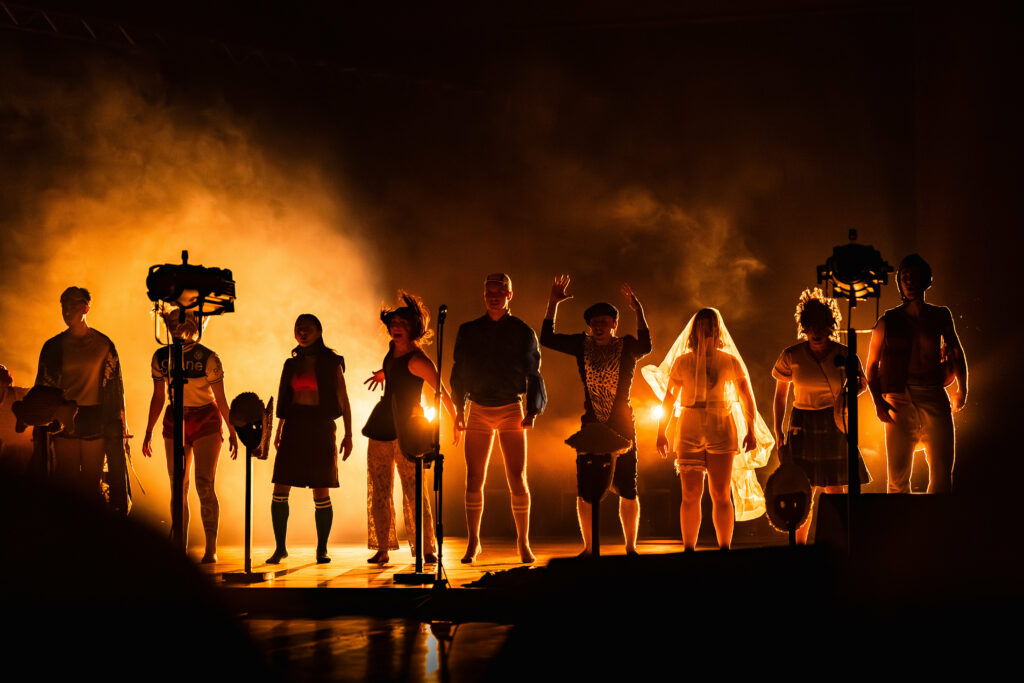 Une rangée de danseurs de la troupe Maxka’roa sur scène à Mauléon-Licharre, silhouettés par une lumière orangée intense et une épaisse fumée de scène. Ils portent des costumes variés et expressifs dans une ambiance dramatique et énergique.